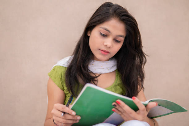 Indian girl student reading and writing on note book. Indian late teen girl student sitting near wall and doing study. She is reading and writing on book in outdoor at day time. She is wearing traditional dress salwar Kameez and Dupatta. beautiful traditional indian girl stock pictures, royalty-free photos & images