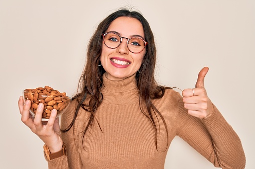 Beautiful woman with blue eyes holding bowl with healthy almonds over white background happy with big smile doing ok sign, thumb up with fingers, excellent sign