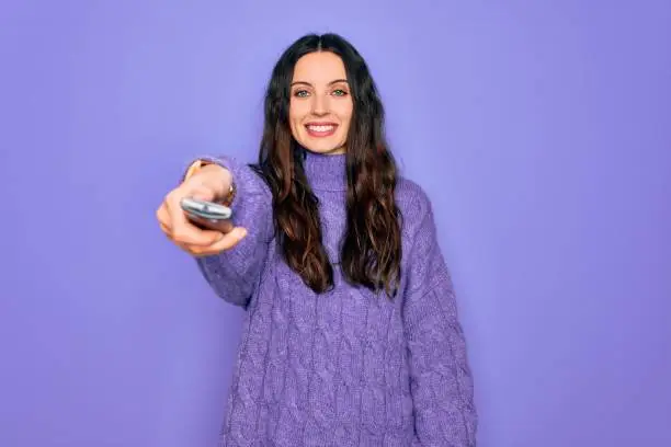 Photo of Young beautiful woman using television remote control over isolated purple background with a happy face standing and smiling with a confident smile showing teeth