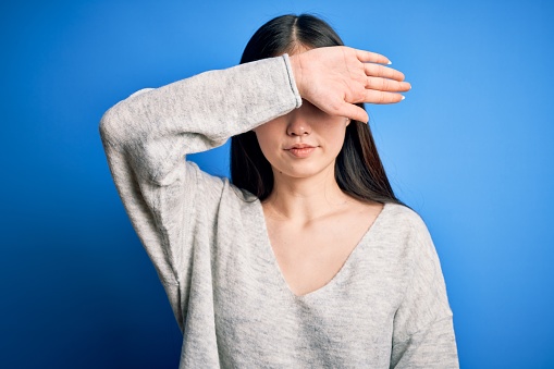Young beautiful asian woman wearing casual sweater standing over blue isolated background covering eyes with arm, looking serious and sad. Sightless, hiding and rejection concept