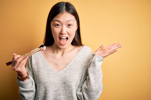 jovem asiática comendo comida japonesa, segurando salmão e arroz maki sushi usando pauzinhos muito feliz e animado, expressão vencedora celebrando vitória gritando com grande sorriso e mãos levantadas - chopsticks human hand women isolated - fotografias e filmes do acervo