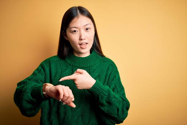 giovane bella donna asiatica che indossa maglione verde invernale su sfondo giallo isolato in fretta indicando il tempo di guardare, impazienza, sconvolto e arrabbiato per il ritardo della scadenza - checking the time women impatient wristwatch foto e immagini stock