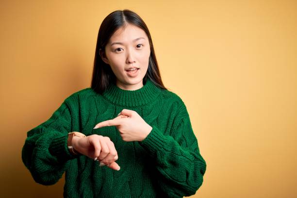 молодая красивая азиатская женщина носить зеленый зимний свитер на желтом изолированном фоне в спешке указывая, чтобы посмотреть время, не - checking the time women impatient wristwatch стоковые фото и изображения