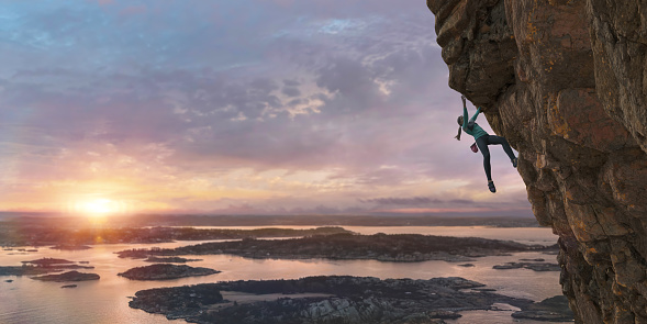 A young female free climber dressed in leggings, long sleeved climbing top, climbing shoes and chalk bag high up on a steep sheer rock face. The climber is in a coastal location as the morning sun rises over water and small islands. With selective focus on the climber.