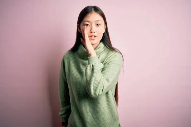 Photo of Young beautiful asian woman wearing green winter sweater over pink solated background hand on mouth telling secret rumor, whispering malicious talk conversation