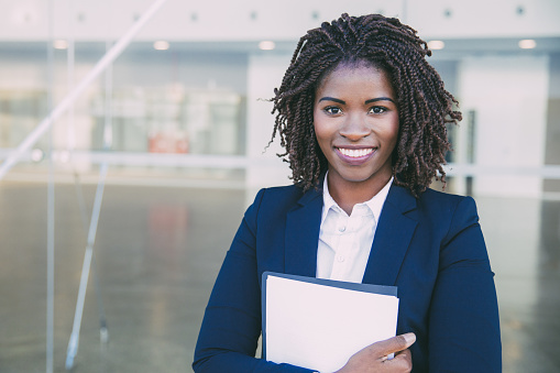 Happy friendly legal expert posing outside. Young black business woman standing at glass wall, holding documents, looking at camera, smiling. Legal expert concept