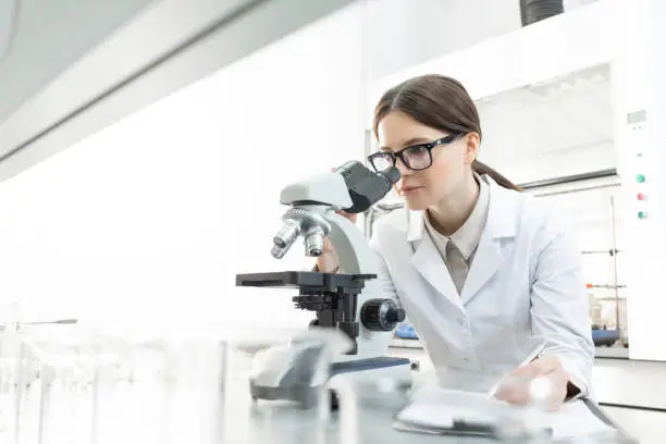 Serious female researcher in whitecoat looking in microscope by workplace while studying features of covid19 in scientific laboratory