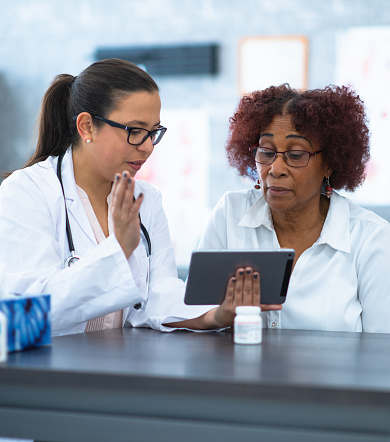 An elderly African American woman wearing glasses sits next to a female doctor.  The female doctor is wearing glasses, a white lab coat and has a stethoscope around her neck.  They are discussing the woman's medical chart