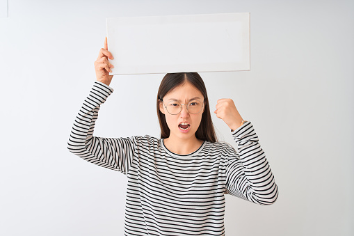 Young chinese woman wearing glasses holding banner over isolated white background annoyed and frustrated shouting with anger, crazy and yelling with raised hand, anger concept