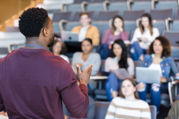 Male college professor gestures during lecture An African American male college professor gestures while giving a lecture to a group of college students. lecture hall photos stock pictures, royalty-free photos & images