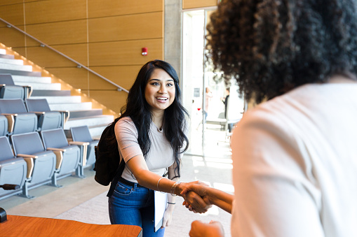 Smiling Hispanic female college student smiles as she shakes hands with a female professor. The student is introducing herself to the professor.