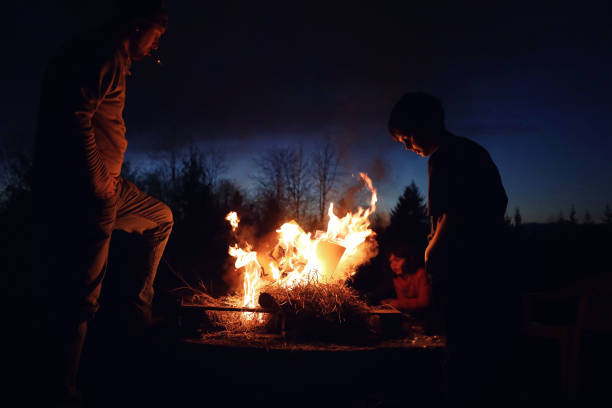 A father and his two young children standing around a fire pit at night looking into the fire.