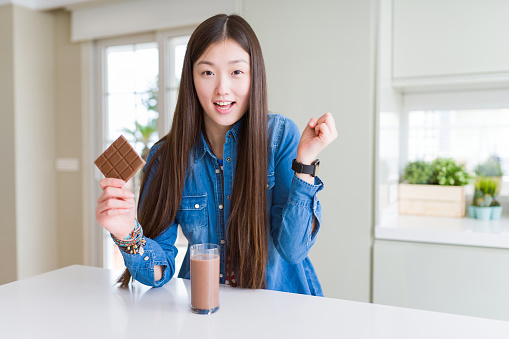 Beautiful Asian woman drinking chocolate milkshake and holding chocolate bar screaming proud and celebrating victory and success very excited, cheering emotion