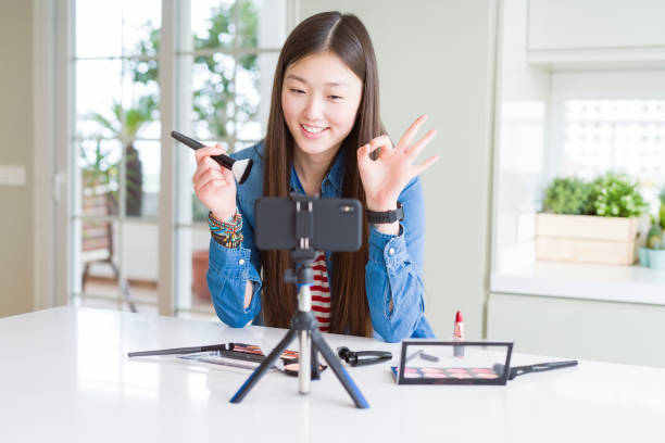 istock Beautiful Asian influencer woman recording make up tutorial using smartphone camera doing ok sign with fingers, excellent symbol 1213742463