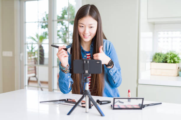 istock Beautiful Asian influencer woman recording make up tutorial using smartphone camera happy with big smile doing ok sign, thumb up with fingers, excellent sign 1213742502