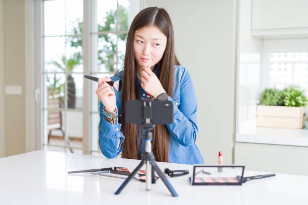 istock Beautiful Asian influencer woman recording make up tutorial using smartphone camera serious face thinking about question, very confused idea 1213742503