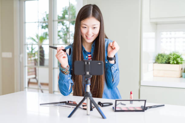 istock Beautiful Asian influencer woman recording make up tutorial using smartphone camera screaming proud and celebrating victory and success very excited, cheering emotion 1213742519