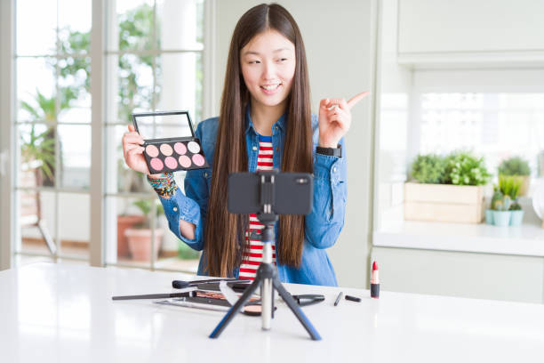 istock Beautiful Asian influencer woman recording make up tutorial using smartphone camera very happy pointing with hand and finger to the side 1213742700