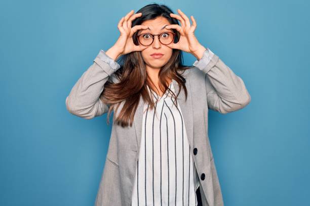 młoda latynoska kobieta biznesu w okularach stojących nad niebieskim odizolowanym tle próbuje otworzyć oczy palcami, senny i zmęczony na poranne zmęczenie - wide awake zdjęcia i obrazy z banku zdjęć