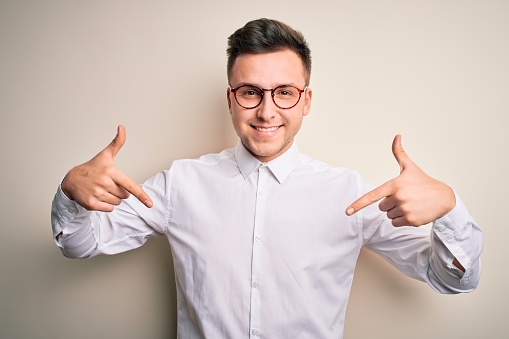 Young handsome business mas wearing glasses and elegant shirt over isolated background looking confident with smile on face, pointing oneself with fingers proud and happy.