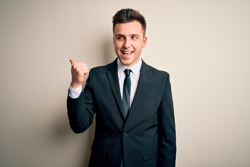 Young handsome business man wearing elegant suit and tie over isolated background smiling with happy face looking and pointing to the side with thumb up.