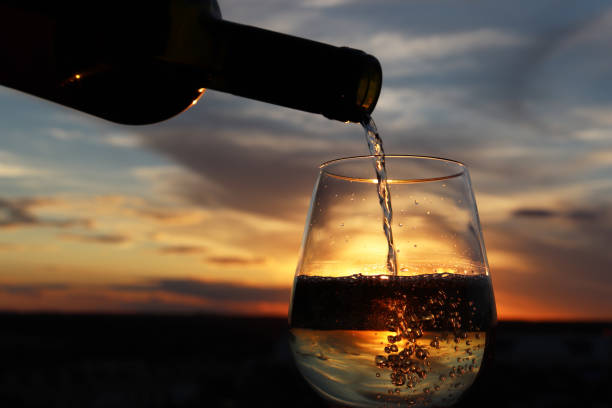 White wine pouring from a bottle into the glass on beautiful sunset background, orange sun is shining through the jet