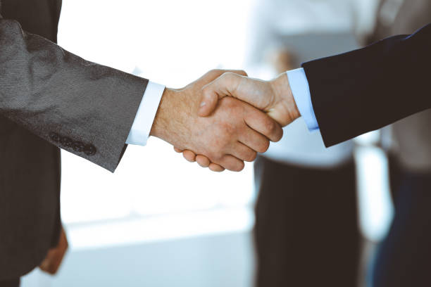istock Business people shaking hands while standing with colleagues after meeting or negotiation, close-up. Group of unknown businessmen and women in modern office. Teamwork, partnership and handshake concept, toned picture 1213752385
