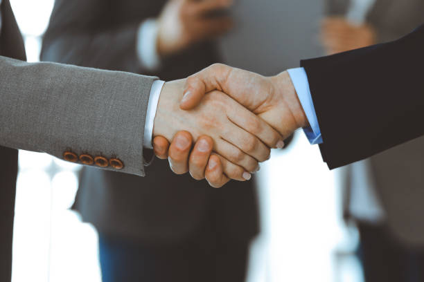 istock Business people shaking hands while standing with colleagues after meeting or negotiation, close-up. Group of unknown businessmen and women in modern office. Teamwork, partnership and handshake concept, toned picture 1213752474