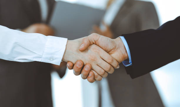istock Business people shaking hands while standing with colleagues after meeting or negotiation, close-up. Group of unknown businessmen and women in modern office. Teamwork, partnership and handshake concept, toned picture 1213752490