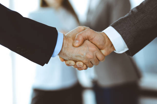 istock Business people shaking hands while standing with colleagues after meeting or negotiation, close-up. Group of unknown businessmen and women in modern office. Teamwork, partnership and handshake concept, toned picture 1213752552