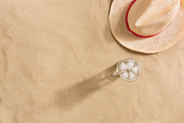 istock Summer vacation composition. Hat and glass of water with ice on sand background. Harsh light with shadows. Summer background. 1213754875