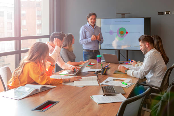 A Manager Dressed In Casual Clothes, Makes A Presentation To His Staff On Business Performace Using A Projector A Large Flat Screen Monitor In The Office Conference Room. stock photo
