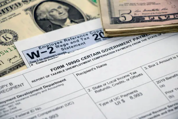 Closeup of a Form 1099G Certain Government Payments for unemployment benefits atop a W2 form and dollar bill, with stack of $5 bills.