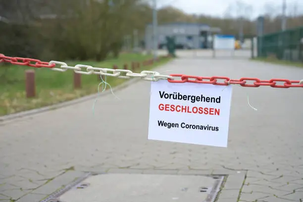 Red white chain barrier in front of a company and sign with German text Vorübergehend Geschlossen, wegen Coronavirus, meaning Temporarily Closed due to Coronavirus.  Countrywide pandemic lock down, copy space, selected focus