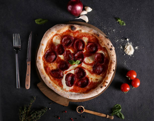 Tasty pepperoni pizza and cooking ingredients tomatoes basil on black concrete background. Top view of hot pepperoni pizza. With copy space for text. Flat lay stock photo