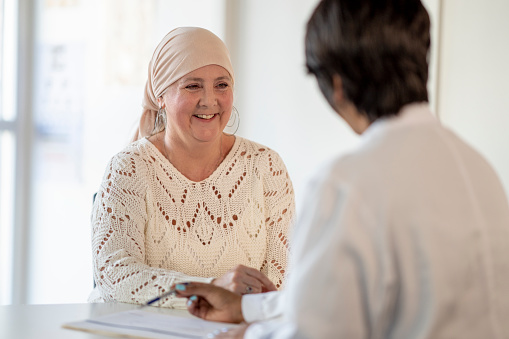 A senior woman with cancer visits the doctor to talk about her treatment plan.  She is dressed casually in a sweater and has a head scarf on.