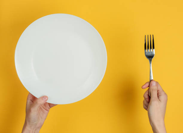istock Minimalistic food concept. Female hands holding a fork and an empty white plate on a yellow background. Top view. Flat lay 1213800504