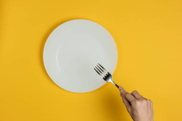 istock Minimalistic food concept. Female hands holding a fork and an empty white plate on a yellow background. Top view. Flat lay 1213800530