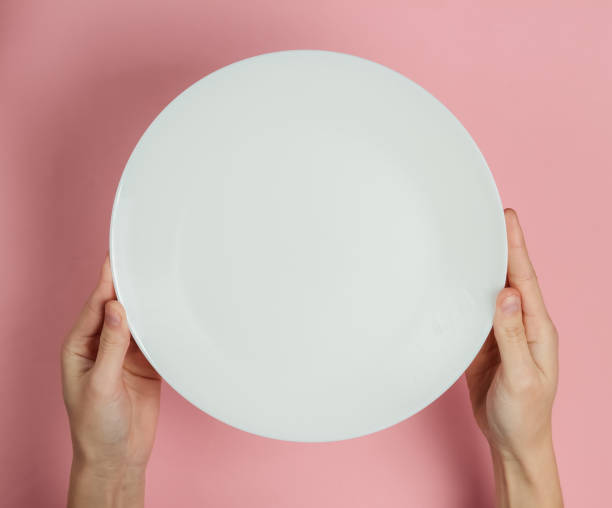istock Female hand holds empty white plate on a pink pastel background. Top view. 1213800645