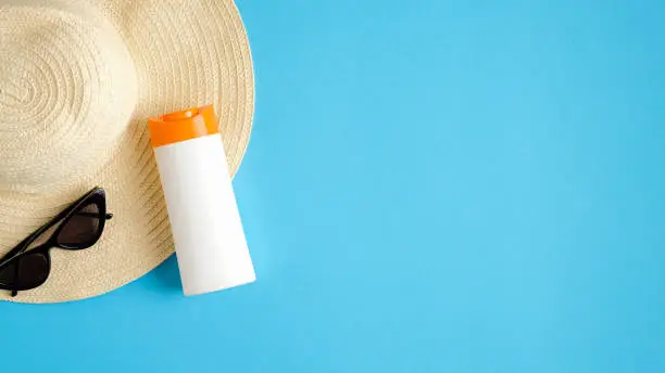 Photo of Blank suntan cream bottle, beach hat and sunglasses on blue background. Sun protection on summer vacation concept