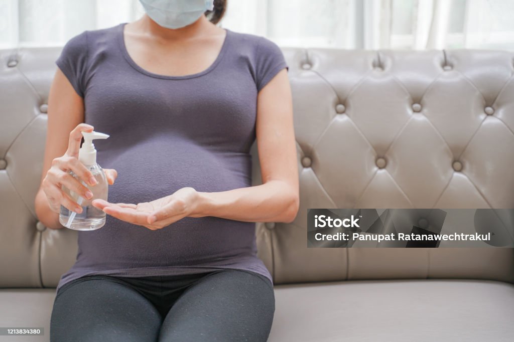 Een zwangere vrouw draagt een chirurgisch masker is handen wassen door alcohol gel op de bank. Bescherm een COVID-19 (Coronavirus), PM 2.5 en voorkom infectie aan het foetusconcept. - Royalty-free Zwanger Stockfoto