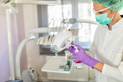 Mature adult female healthcare and medical worker cleaning and disinfecting equipment, surfaces and tools in dental office. She is using alcohol and antiseptic wipes which is necessary after corona virus outbreak. She wears protective mask, gloves and eye wear.