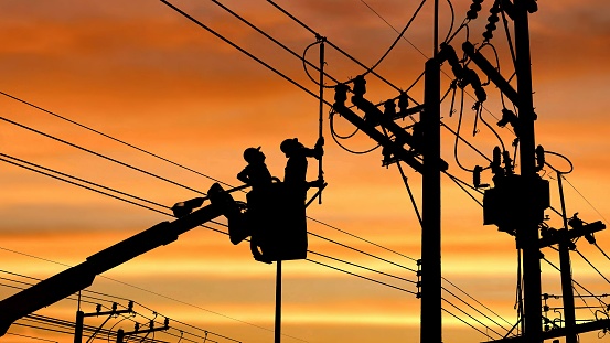 Silhouette two electricians with disconnect stick tool on crane truck are working to install electrical transmission on power pole with blurred sunrise sky background in technology concept