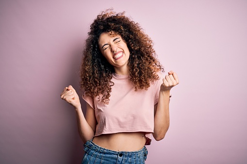 Young beautiful woman with curly hair and piercing wearing casual pink t-shirt very happy and excited doing winner gesture with arms raised, smiling and screaming for success. Celebration concept.