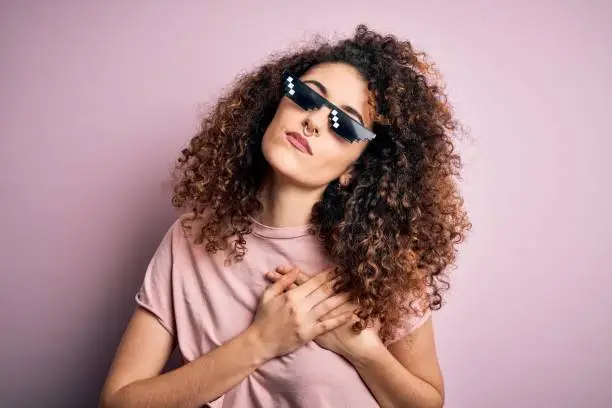 Young beautiful woman with curly hair and piercing wearing funny thug life sunglasses smiling with hands on chest with closed eyes and grateful gesture on face. Health concept.