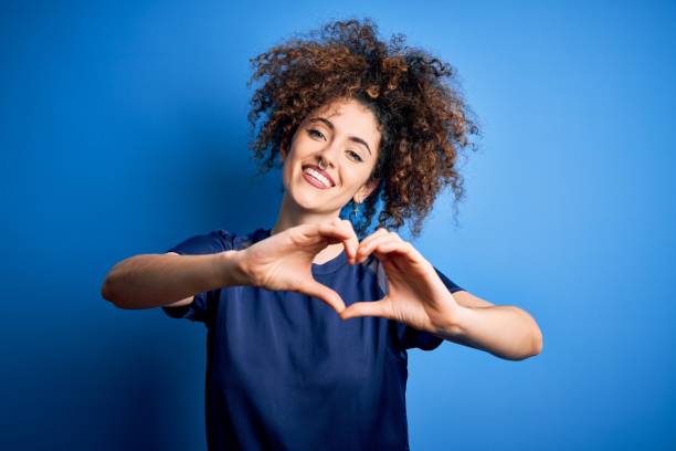 Young beautiful woman with curly hair and piercing wearing casual blue t-shirt smiling in love doing heart symbol shape with hands. Romantic concept. Young beautiful woman with curly hair and piercing wearing casual blue t-shirt smiling in love doing heart symbol shape with hands. Romantic concept. blue t shirt stock pictures, royalty-free photos & images