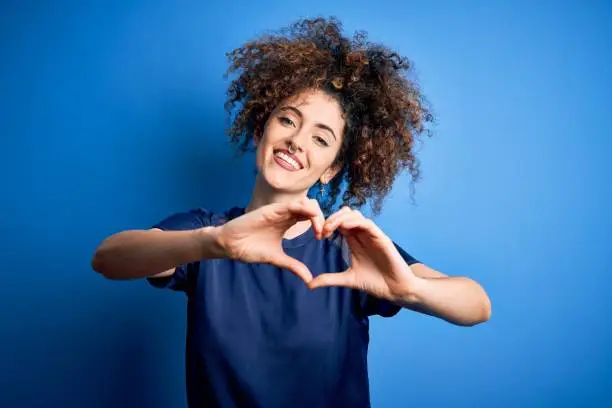 Photo of Young beautiful woman with curly hair and piercing wearing casual blue t-shirt smiling in love doing heart symbol shape with hands. Romantic concept.