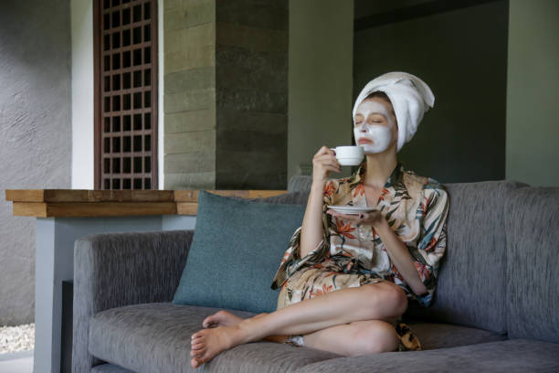 joven sentada en casa con máscara de belleza facial mientras disfruta de una taza de café. acogedor tiempo en casa. tiempo propio y concepto de spa en casa. brote de coronavirus y concepto de auto-cuarentena. - bathrobe women cup towel fotografías e imágenes de stock