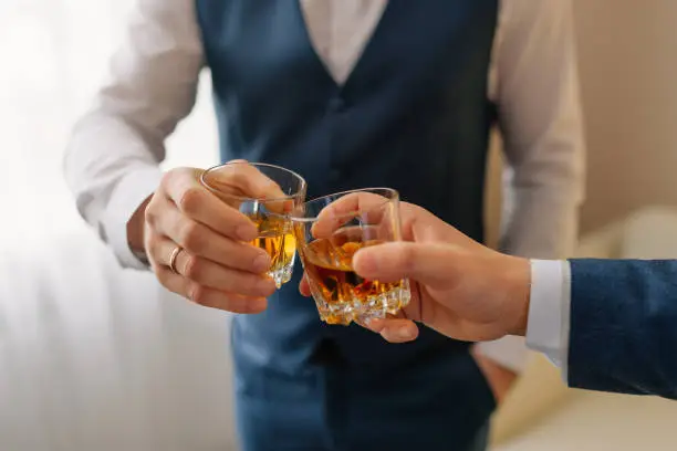 The groom and groomsman drinking a whiskey, celebrating wedding day. Hands of man holding glasses with alcohol and toasting. group of men cheering and clinking with drinks at wedding reception