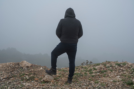 A facing back hoody adult man hands in pocket, standing up the hill and looking at the view in air pollution, foggy weather